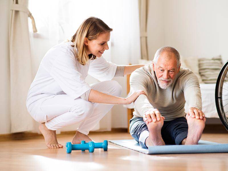 Elderly man stretching on mat and touching his toes while therapist encourages him.