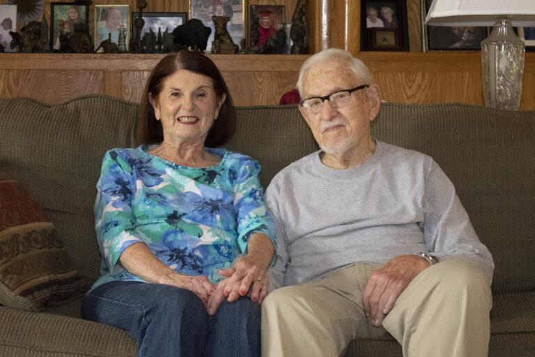 Elderly man and woman on couch holding hands looking at camera and smiling